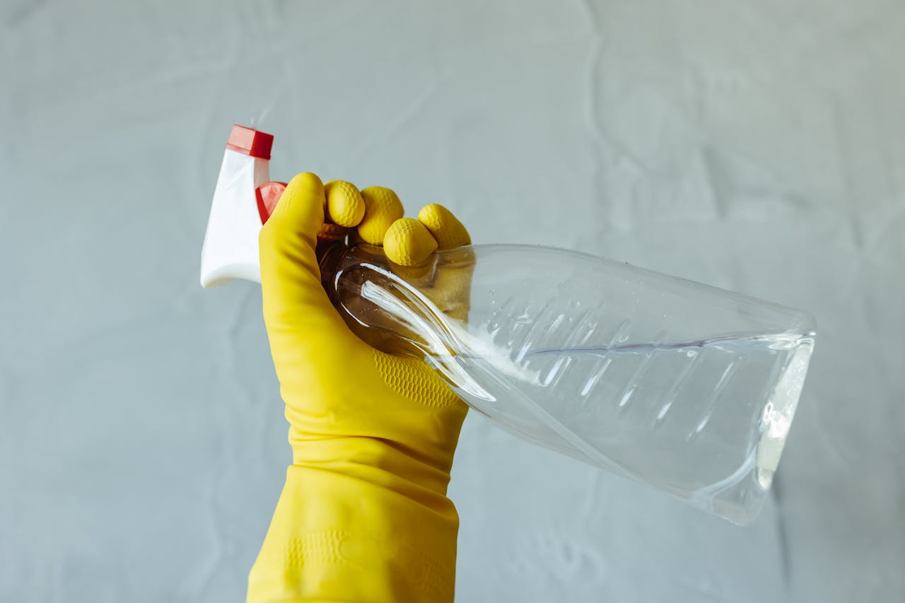 hero-img-02 Close-up of a hand wearing yellow gloves holding a transparent spray bottle, isolated on a plain background.