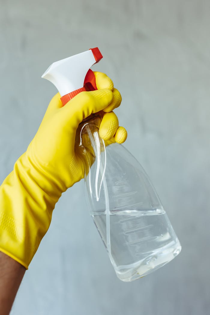 about-01 Yellow gloved hand holding a transparent cleaning spray bottle against a neutral background.
